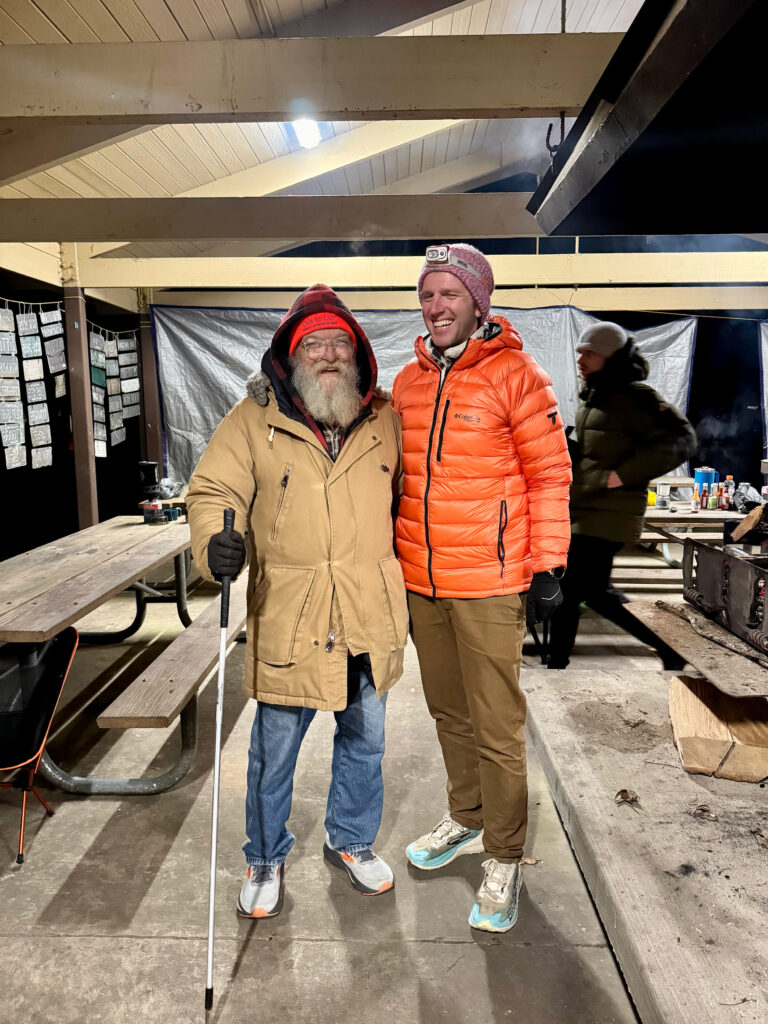 Lazarus Lake, in a plaid hat and tan coat, at a night-time race camp