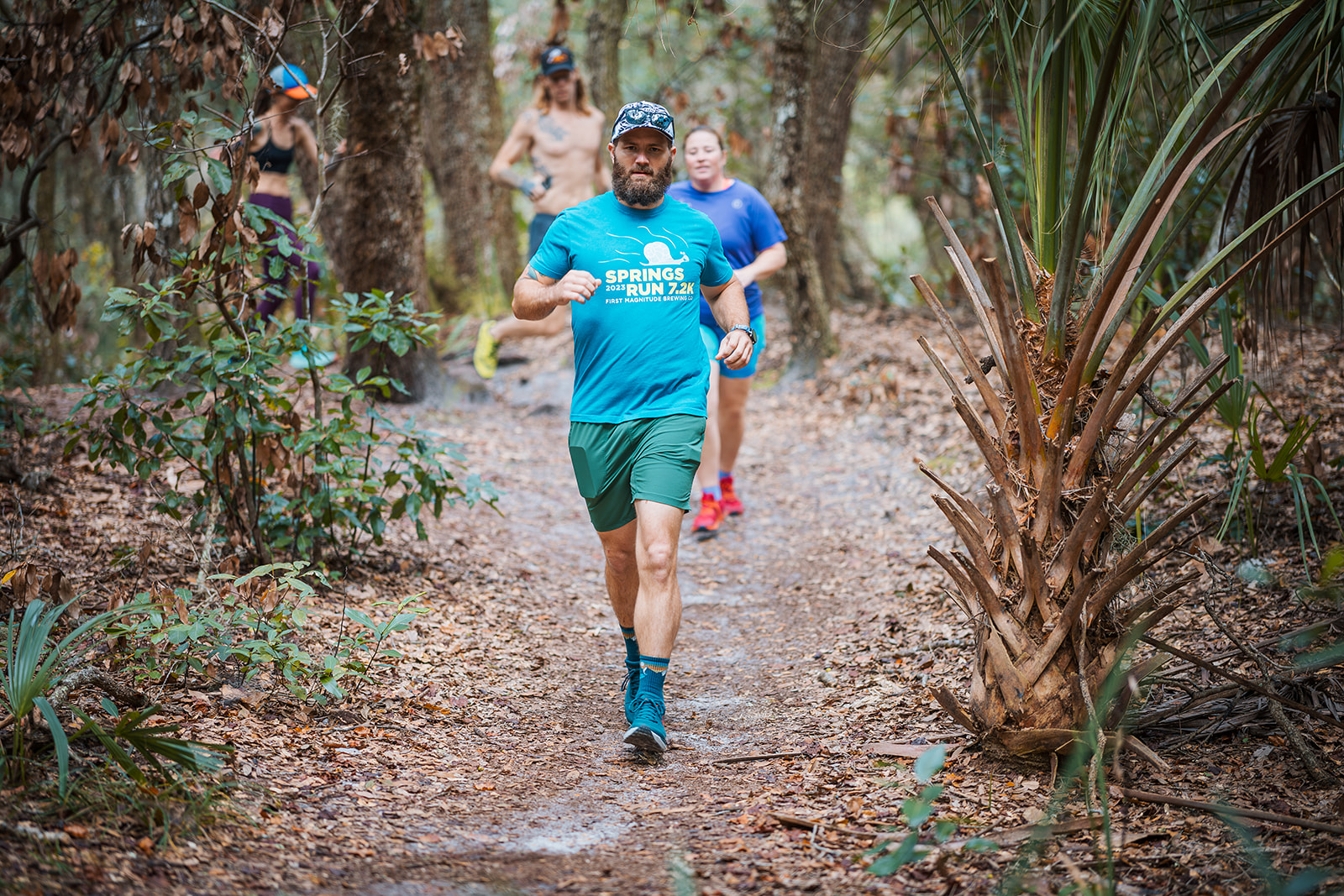 A runner on a leaf covered trail through palmetto and live oak, more runners on the path behind
