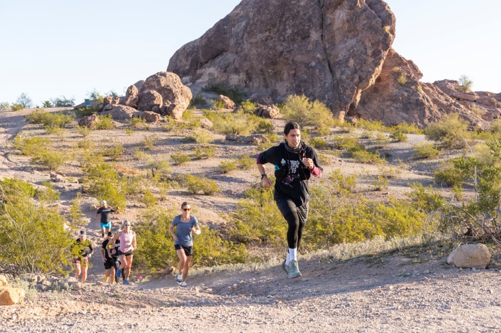 A runner coming down a rocky desert trail with more runners behind, a sandstone outcrop and open sky above