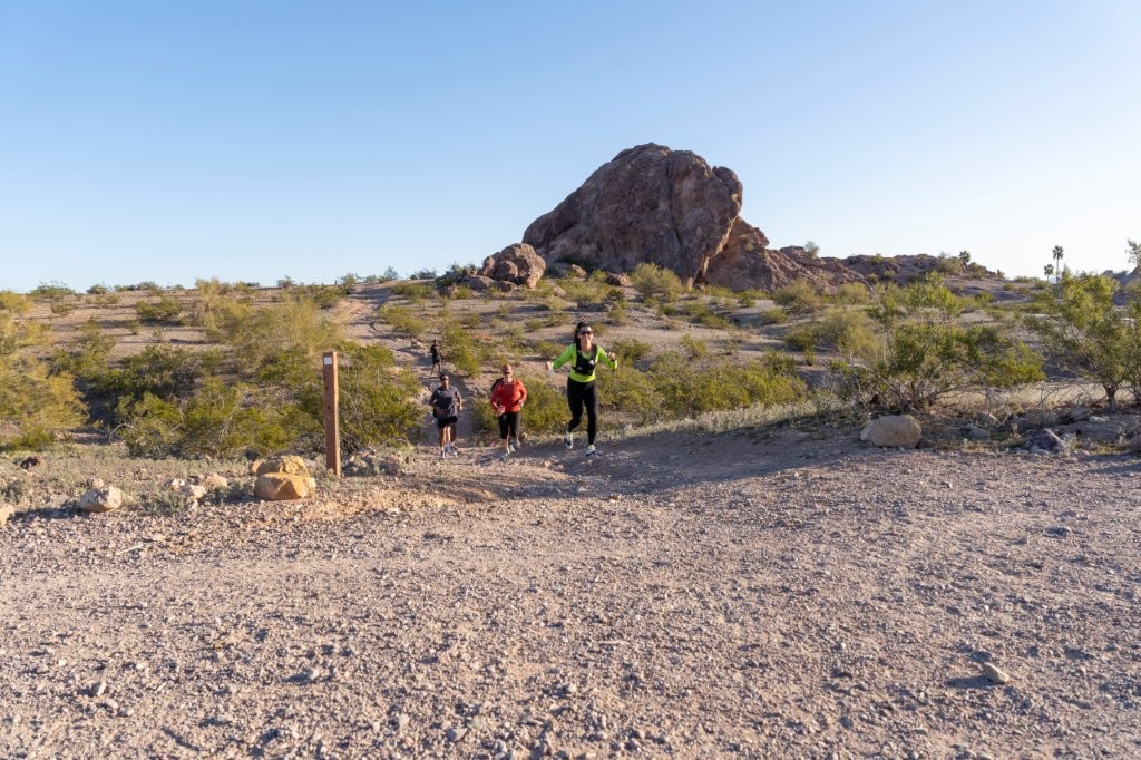 Three runners strung out along a wide gravel trail through desert scrub, a sandstone outcrop behind them