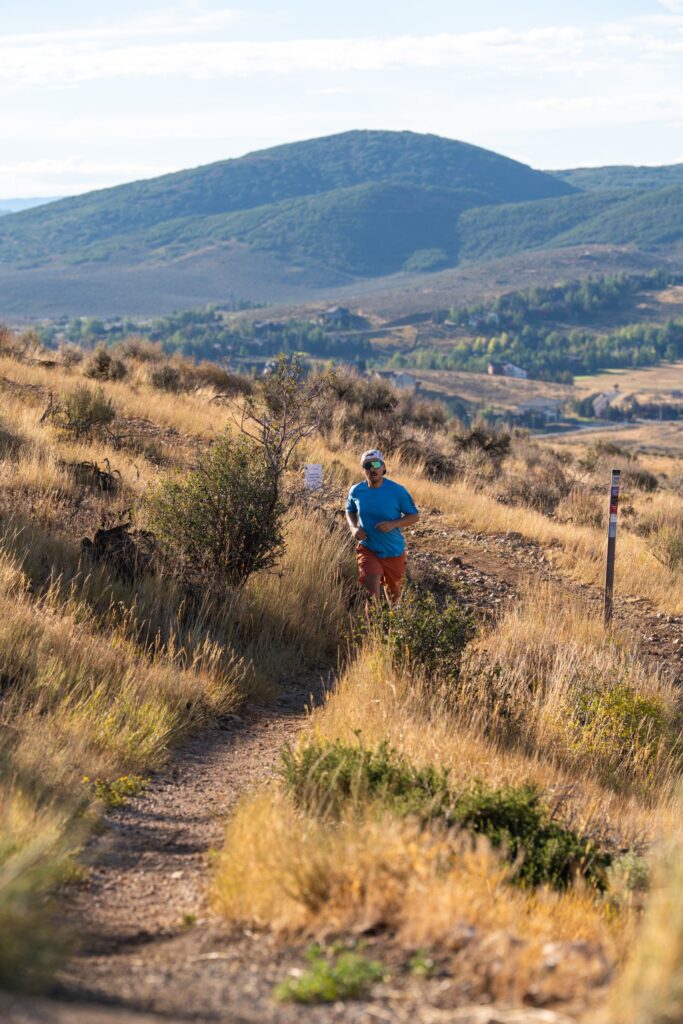 A runner on a dry grass singletrack trail, a green ridge and open sky behind