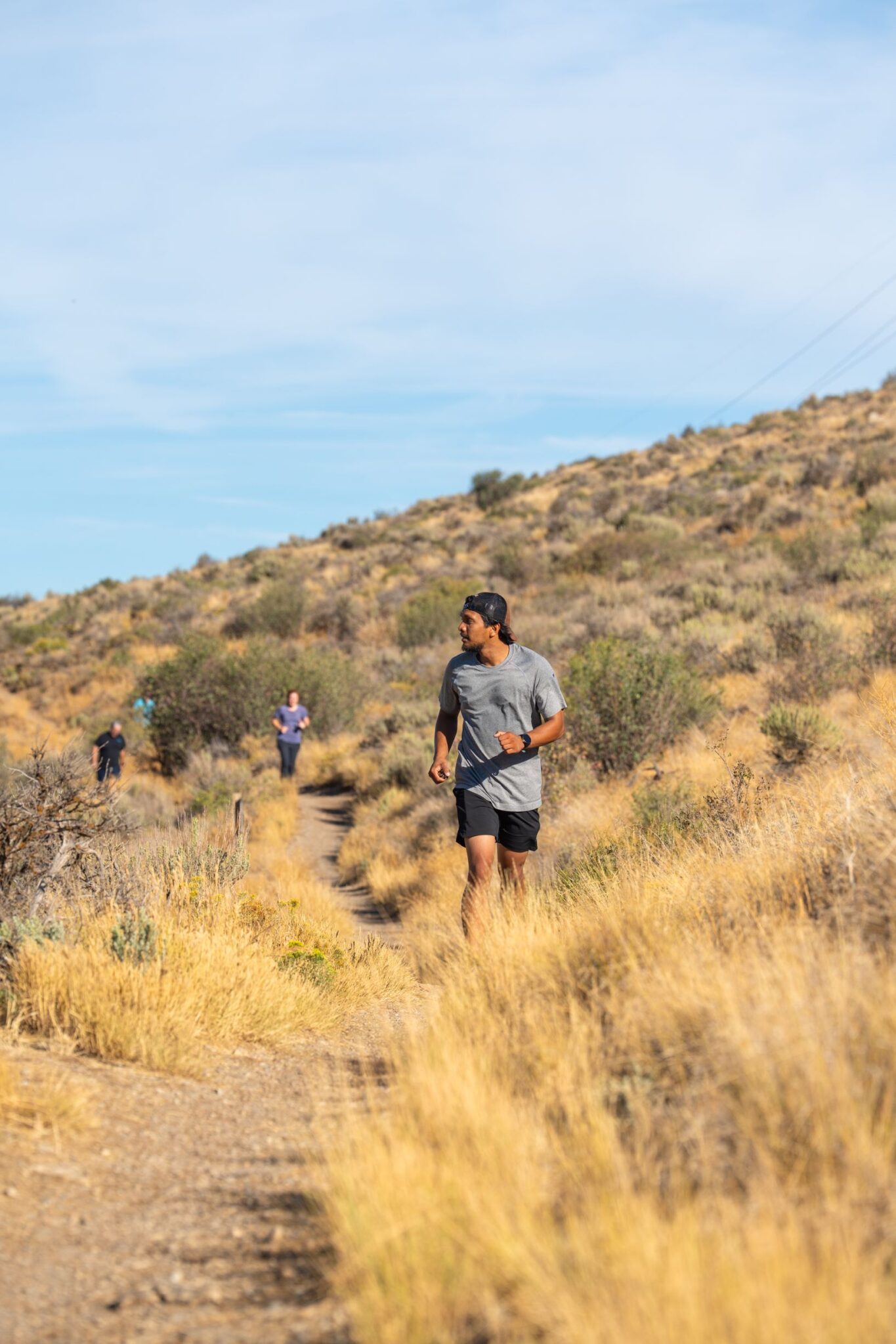 A runner on a dry grass singletrack trail with more runners strung out along the hillside behind, a green ridge and open sky above
