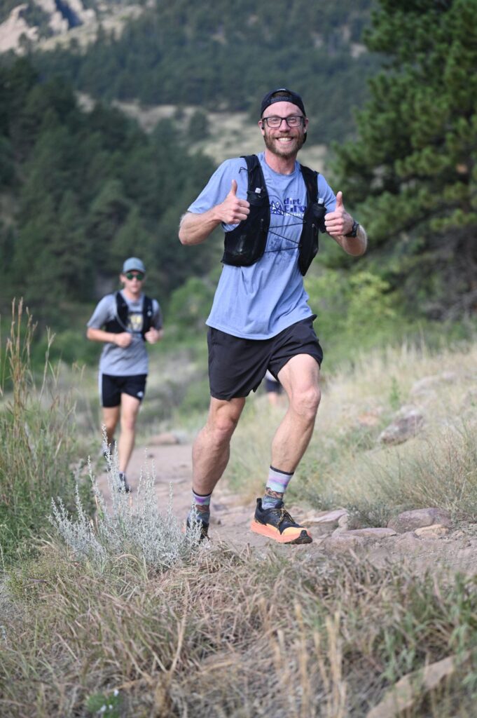 A runner in a Dirt Circus shirt grinning and giving two thumbs up on a rocky trail