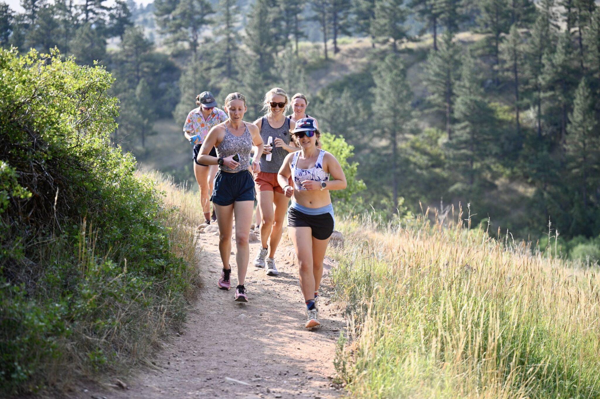 Five runners coming toward the camera on a narrow dirt trail, ponderosa pines and dry golden grass on the hillside behind them