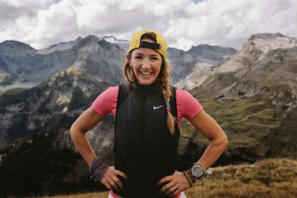 Sally McRae smiling with her hands on her hips on a mountain ridge, peaks and cloud behind her